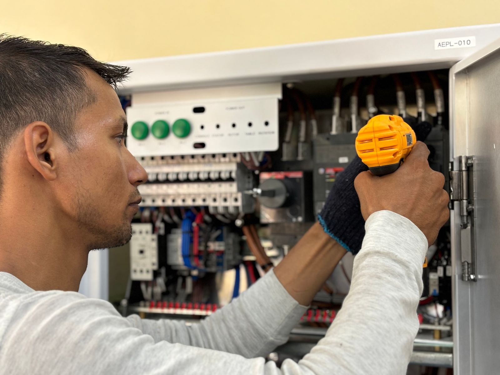 An electrician in a light grey top using a yellow cordless driver at an open electrical control panel